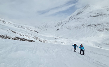 Zwei Wanderer beim Winterwandern im verschneiten Hochtal Juf in Graubünden, umgeben von schneebedeckten Bergen und blauem Himmel.
