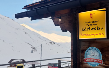 Restaurant Edelweiss in Juf mit gelbem Schild und Blick auf verschneite Alpen im Hintergrund.
